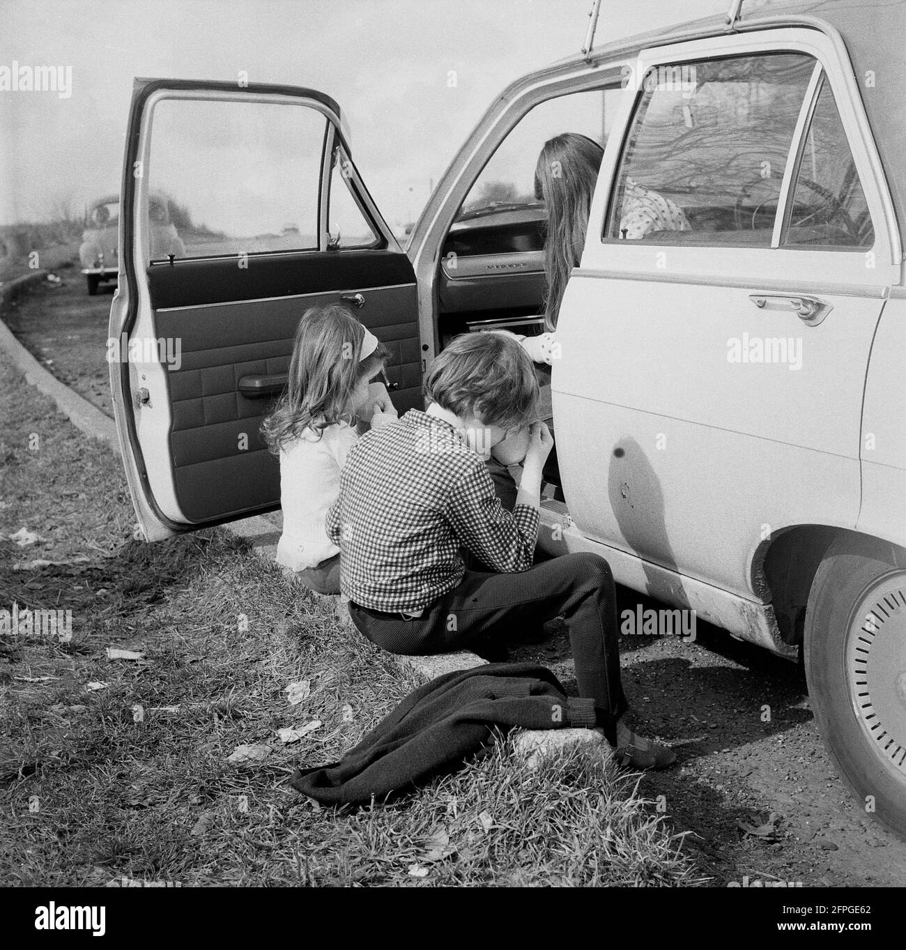 1969, historical, two children sitting on the kerb, having a drink, waiting outside their dad's car, which is parked on a roadside parking area, Wrotham Hill, Essex. England, UK. Stock Photo