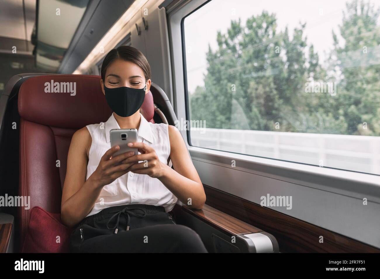 Face mask in public transport during corona virus. Elegant Asian passenger woman sitting in business class of train using mobile phone during travel Stock Photo