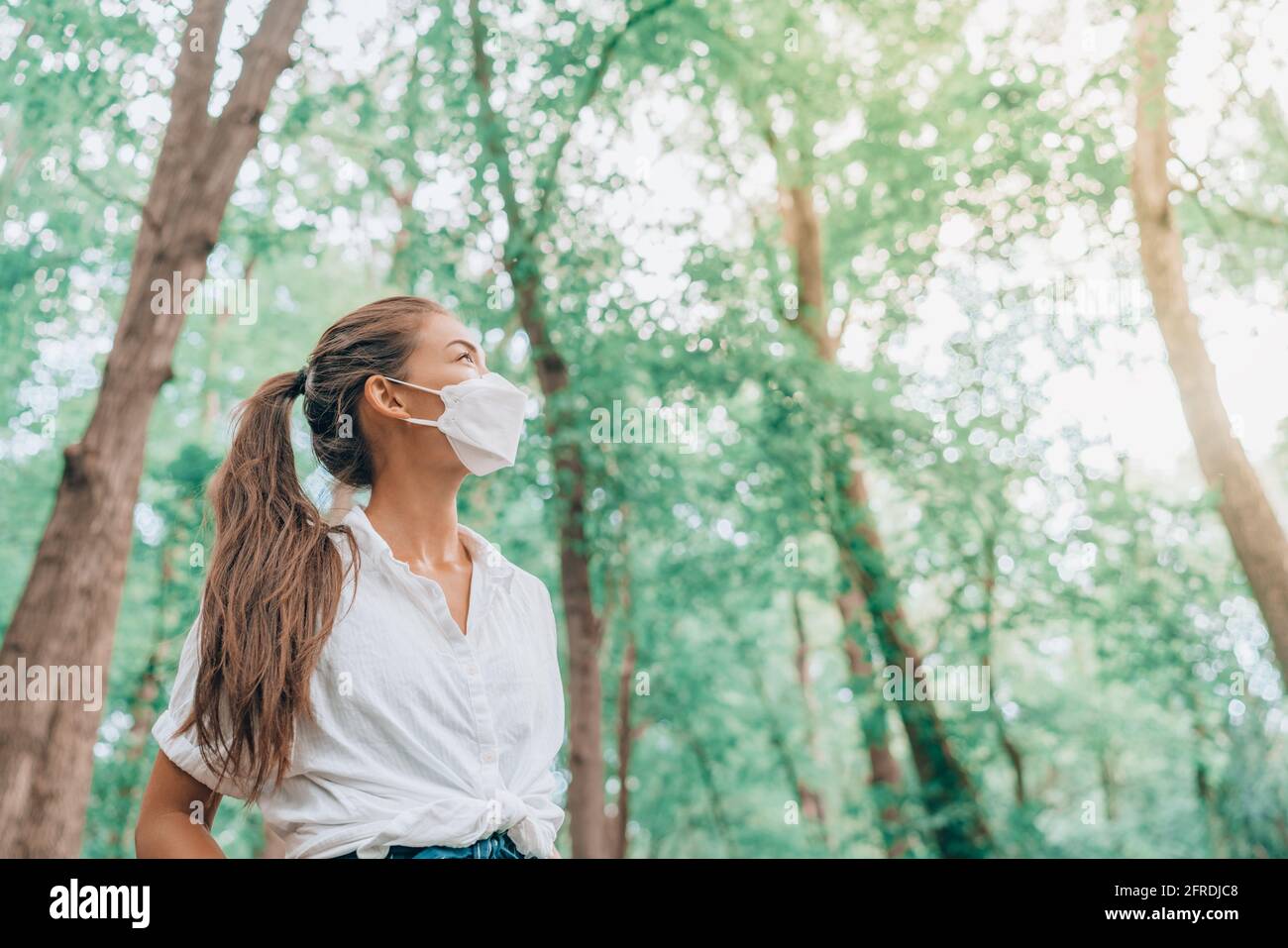 Mask wearing woman looking up to bright future in hope walking in forest. Sustainable fashion fabric in PPE, asian girl lifestyle breathing clean air Stock Photo