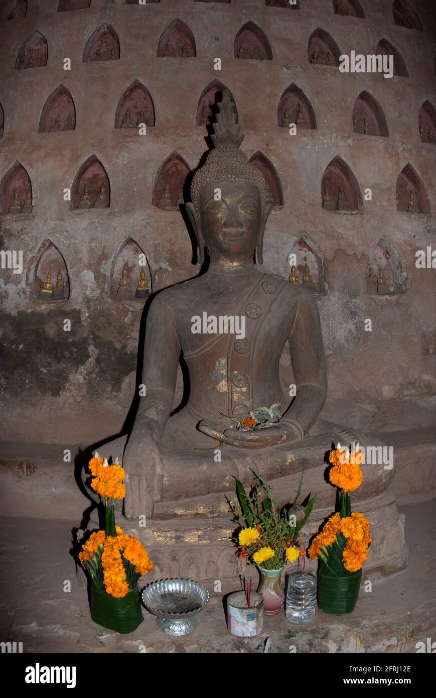 Statue of seated Buddha with flowers, Cloisters with small Buddhas in niches in wall, Wat Si Saket, Vientiane, Lao Stock Photo