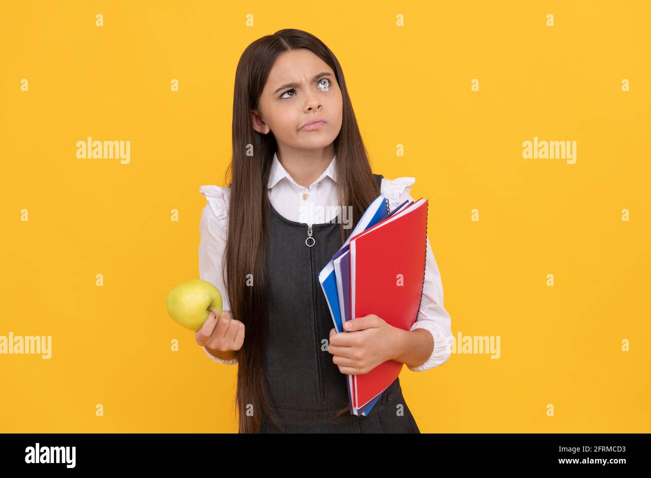 Serious school-aged girl child think holding apple and books yellow background, thinking Stock Photo