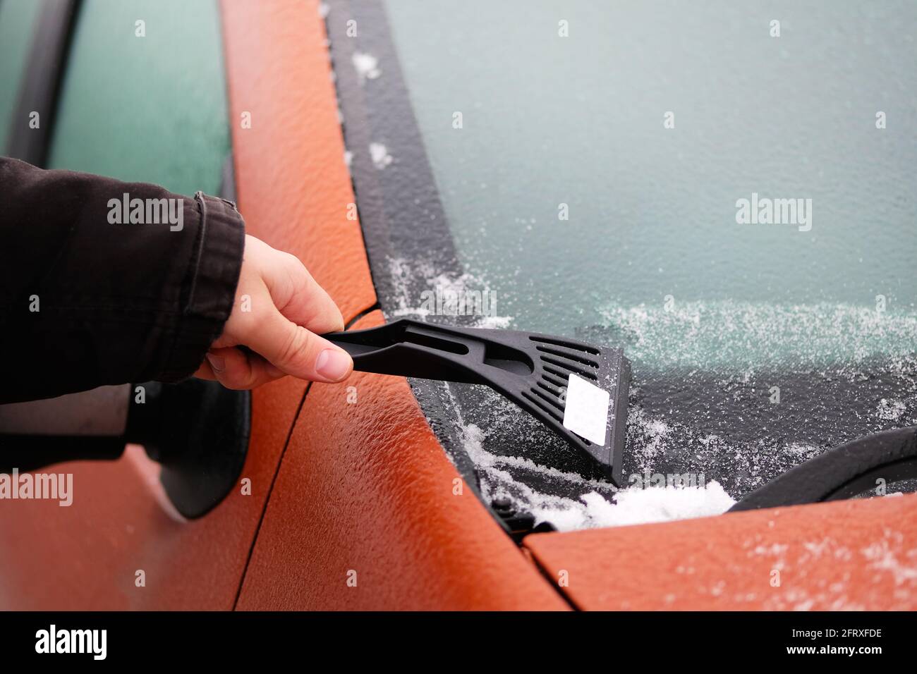 Scraper in mans hand. Man clears snow from icy windshield of car. Cleaning orange automobile window. Stock Photo