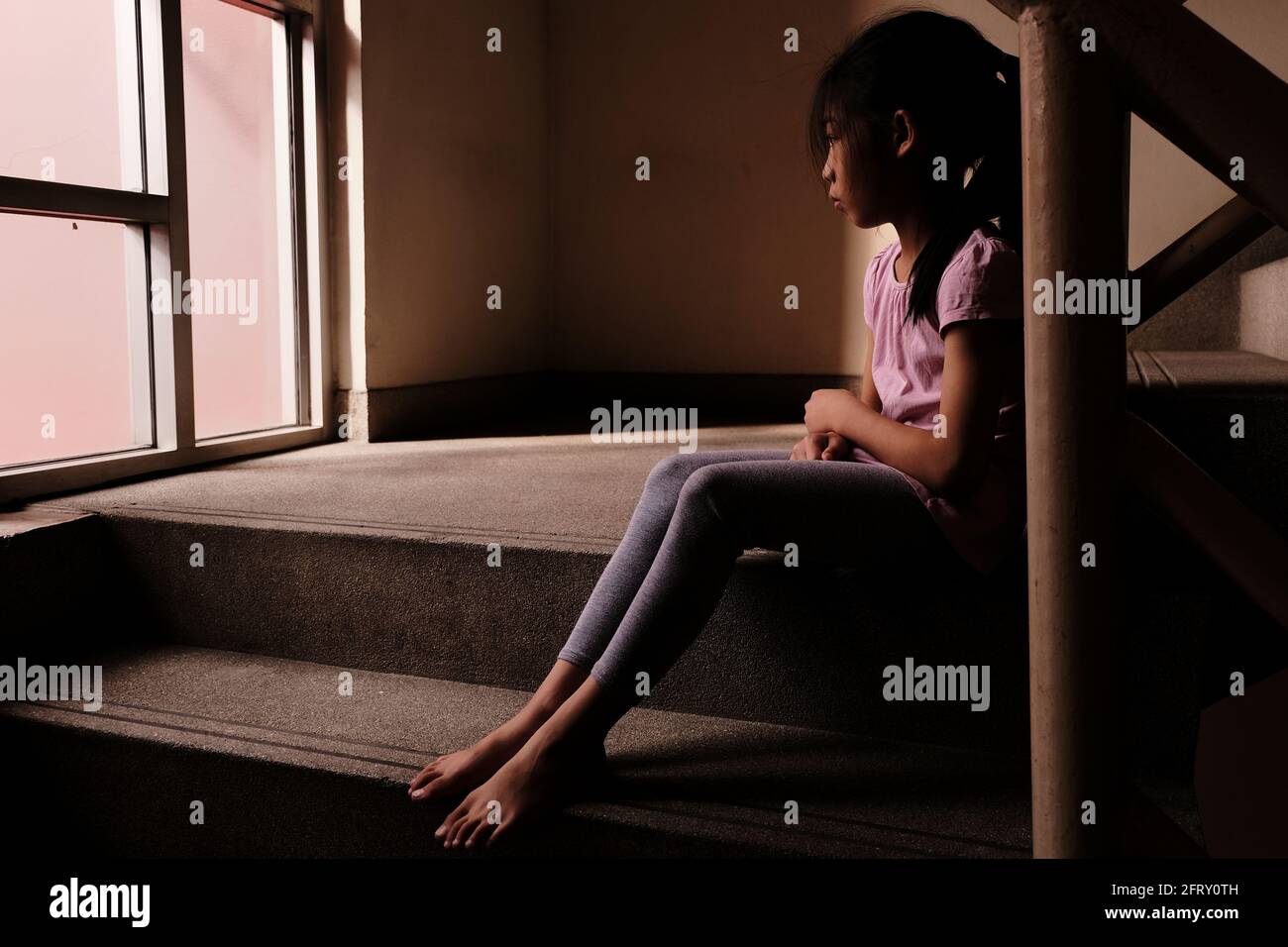 Child abuse concept. A sad and lonely young girl sitting by a stair inside a build with light through glass window in the background. Stock Photo