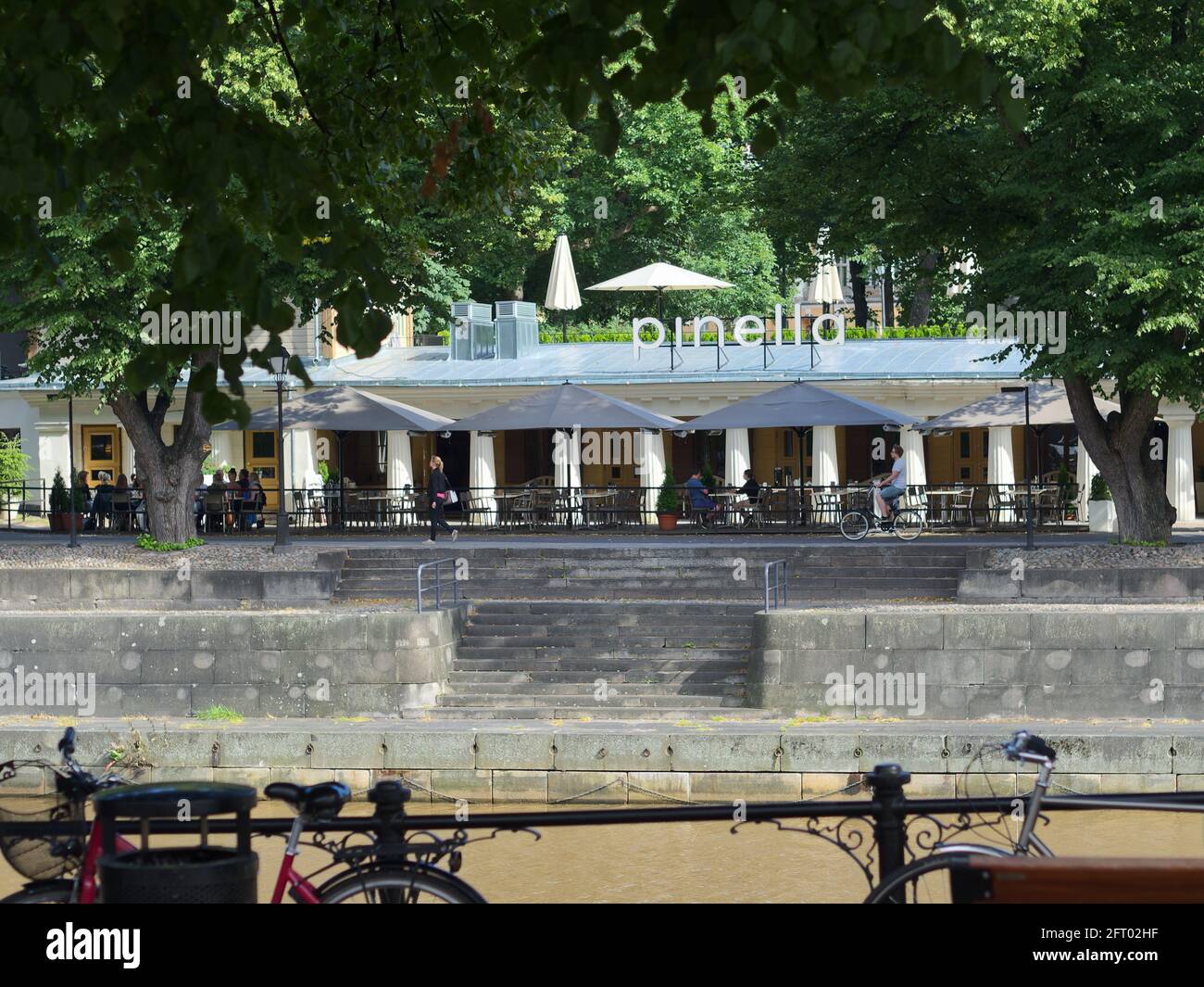 View of restaurant Pinella over the river Aura in Turku, Finland. People are dining outdoors on a warm and sunny summer day. Stock Photo