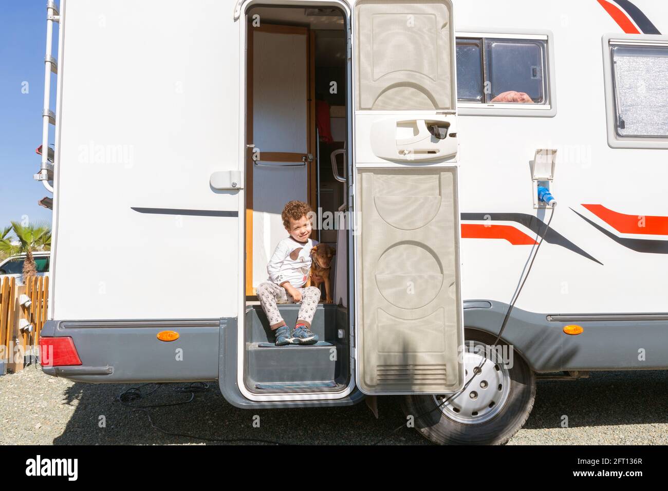 little curly-haired boy smiling sitting in the door of motor home, tenderly holding his pet. Stock Photo