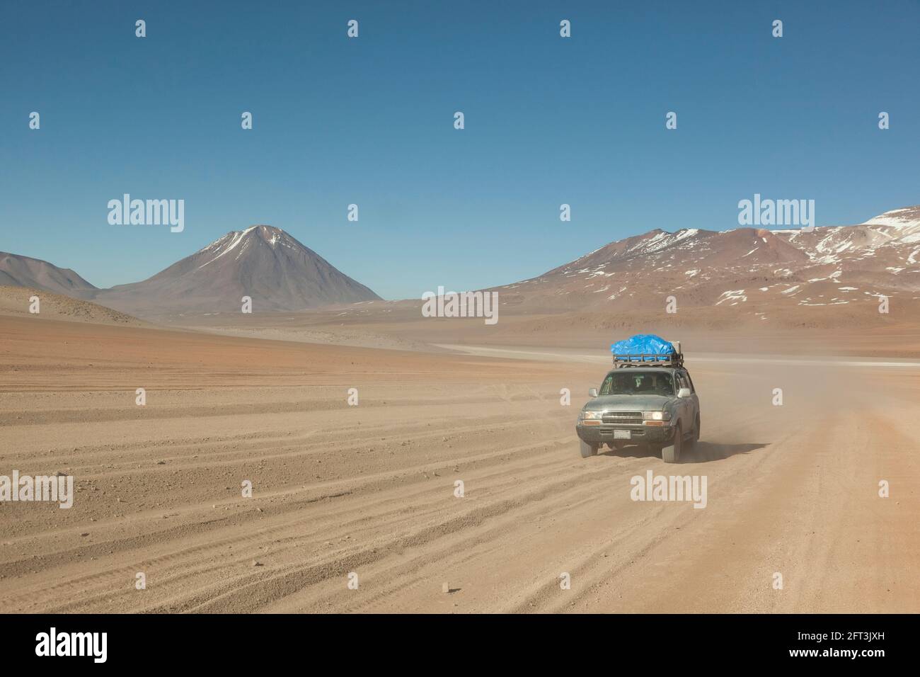 Against a backdrop of volcanoes, a 4x4 off-road vehicle speeds across Bolivia's desert landscape on an overland tourist safari. Stock Photo