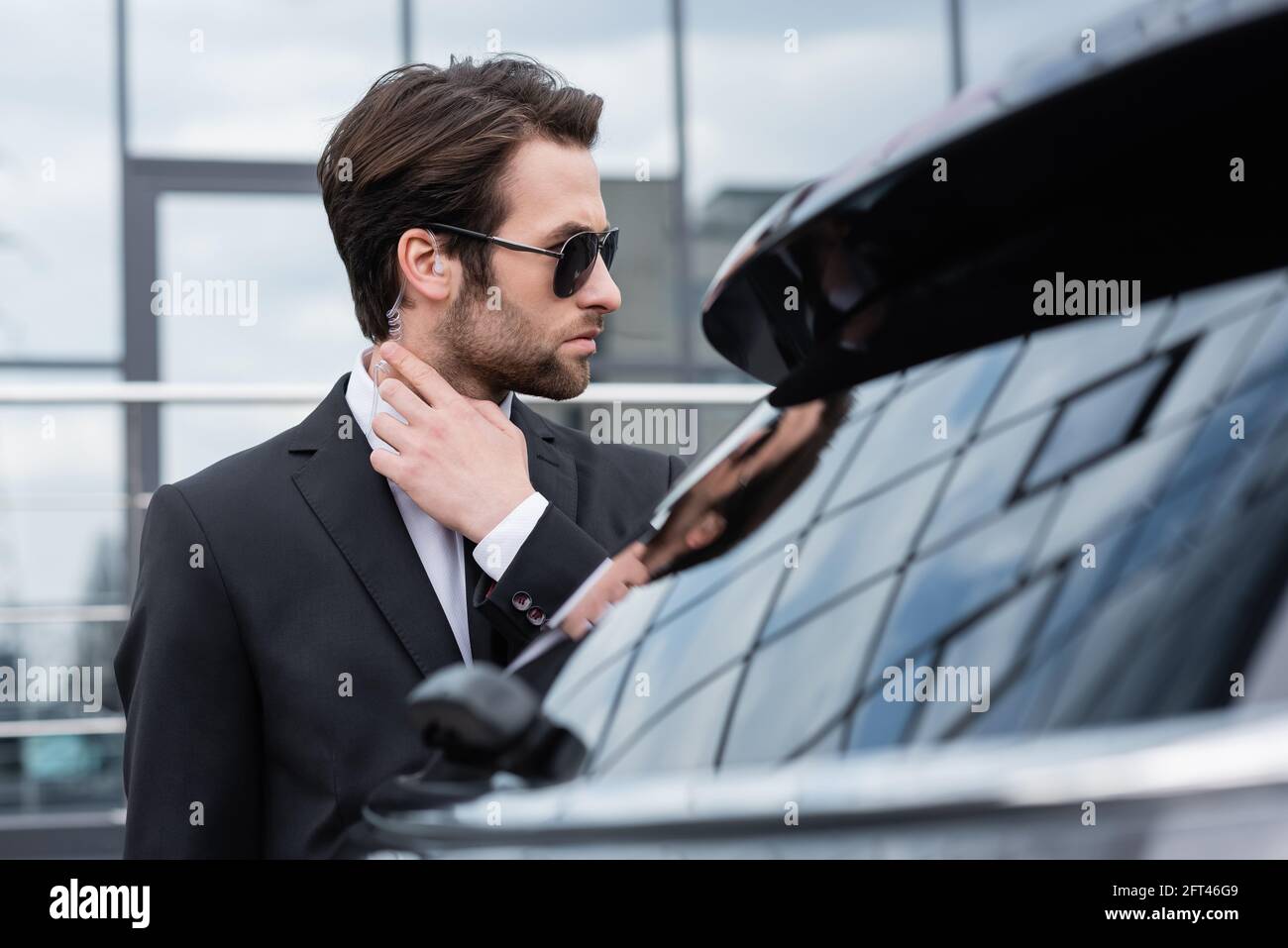 https://c8.alamy.com/comp/2FT46G9/bearded-bodyguard-in-suit-and-sunglasses-adjusting-security-earpiece-near-blurred-car-2FT46G9.jpg