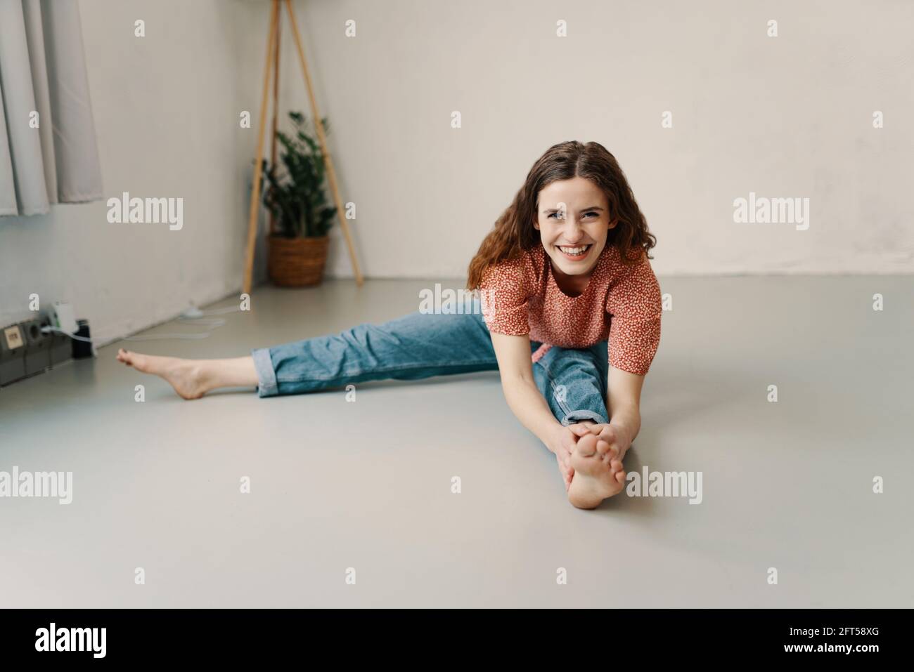 Vivacious young woman working at doing stretching exercises barefoot on the floor at home reaching forwards to grasp her toes as she smiles happily at Stock Photo