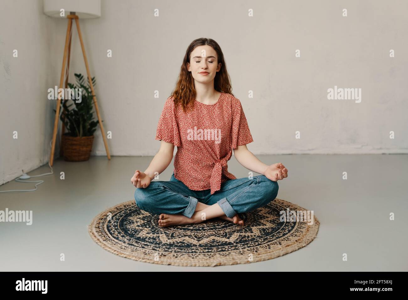 Serene young woman sitting meditating in the lotus pose on a circular mat on the floor at home in a wellness and relaxation concept Stock Photo
