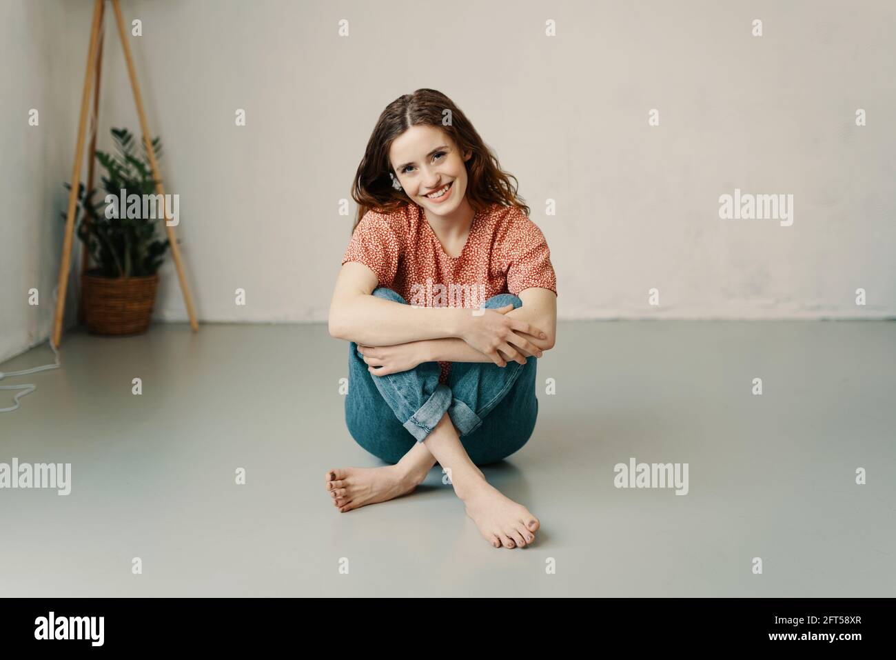 Sweet barefoot young woman with a lovely kind smile sitting on the floor in her jeans relaxing looking at the camera Stock Photo