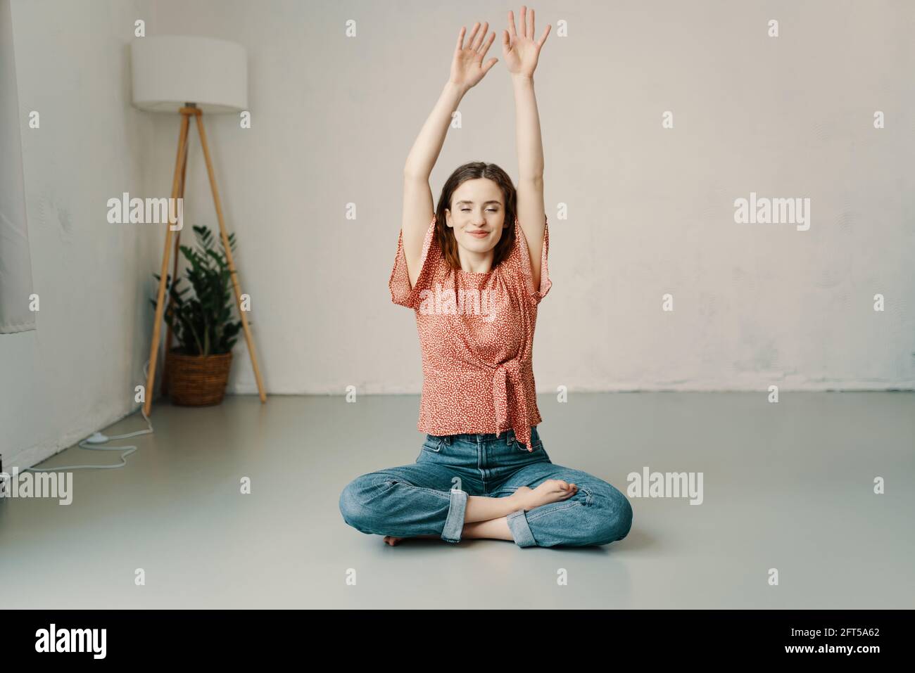 Serene young woman meditating with raised arms, closed eyes and a blisful expression in the lotus pose on the floor at home Stock Photo