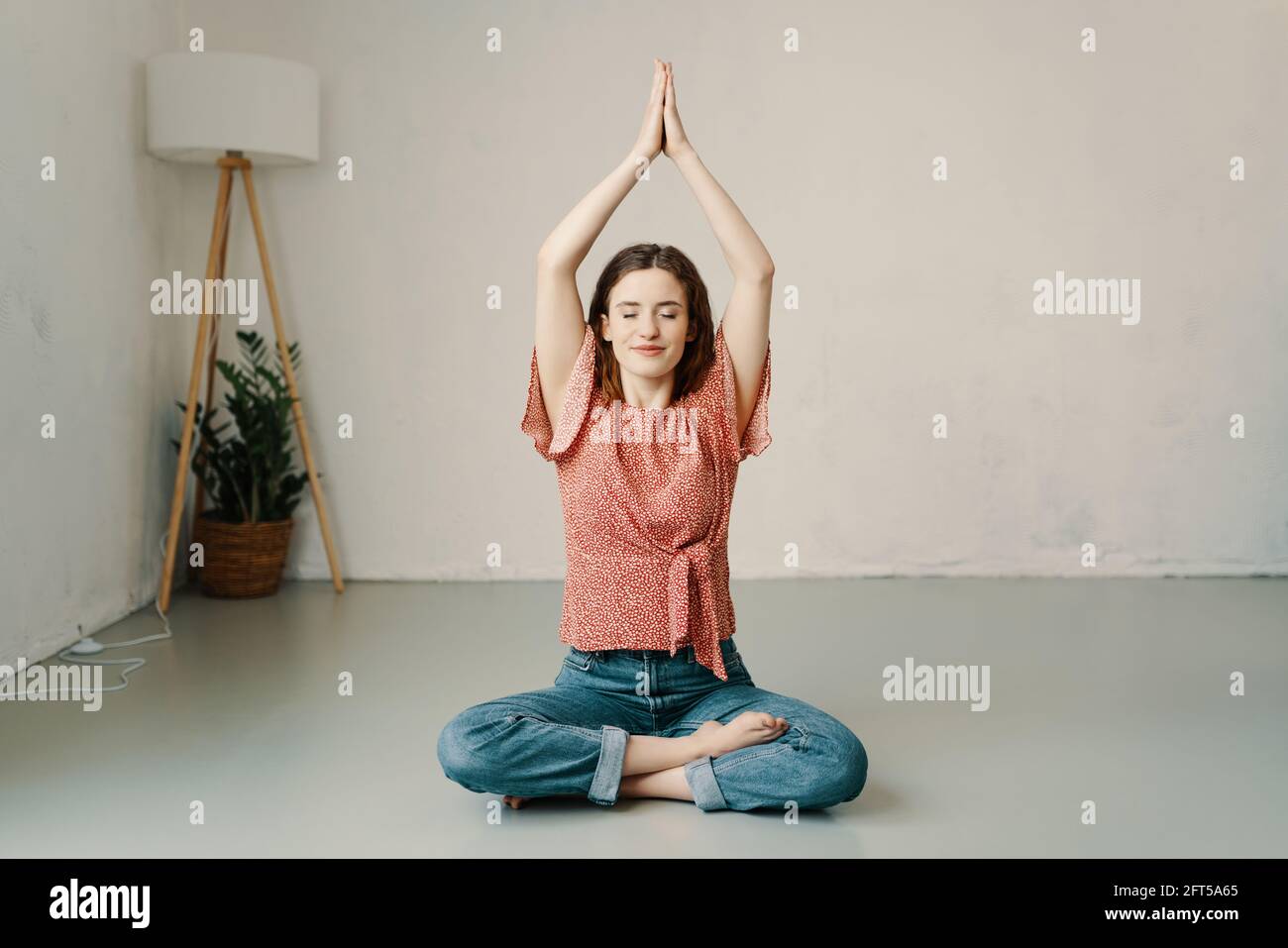 Young woman sitting in the lotus pose meditating on the floor at home with raised hands steepled together and a serene pose with quiet smile and close Stock Photo