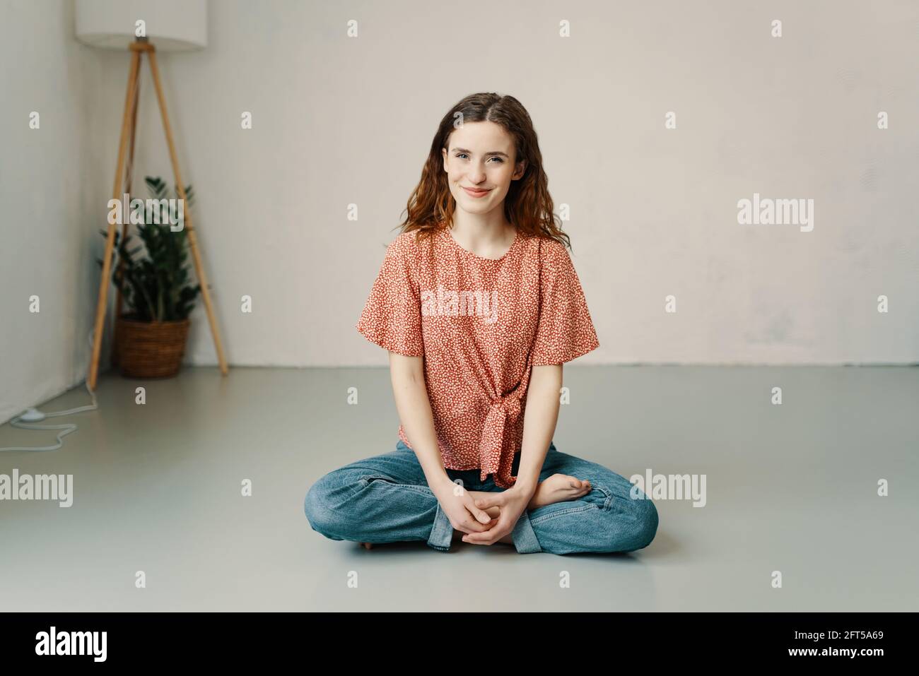 Young woman with a lovely wide warm friendly smile and sweet expression relaxing cross-legged on the floor in the lotus pose with bare feet and copysp Stock Photo