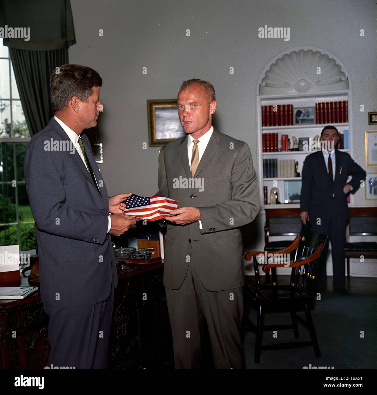 President John F. Kennedy receives a gift of an American flag from astronaut Lieutenant Colonel John H. Glenn, Jr. (right); Lt. Col. Glenn carried the flag in his space suit during his orbital flight aboard Mercury-Atlas 6, also known as Friendship 7. Special Assistant to the President, Kenneth P. O'Donnell, stands in the background. Oval Office, White House, Washington, D.C. Stock Photo