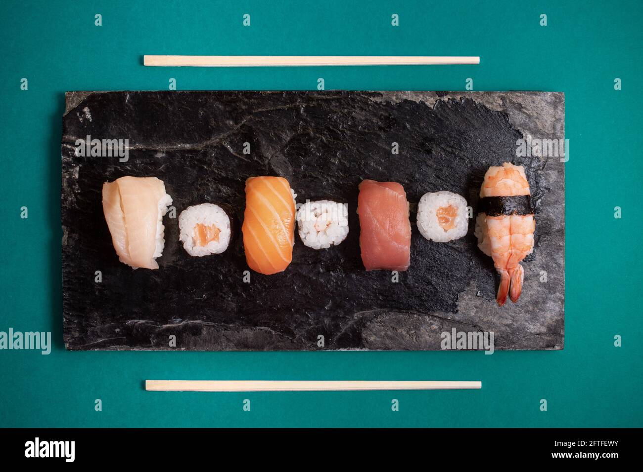Horizontal photograph of sushi on a black plate with textures and sushi sticks on the sides. Turquoise background. Stock Photo