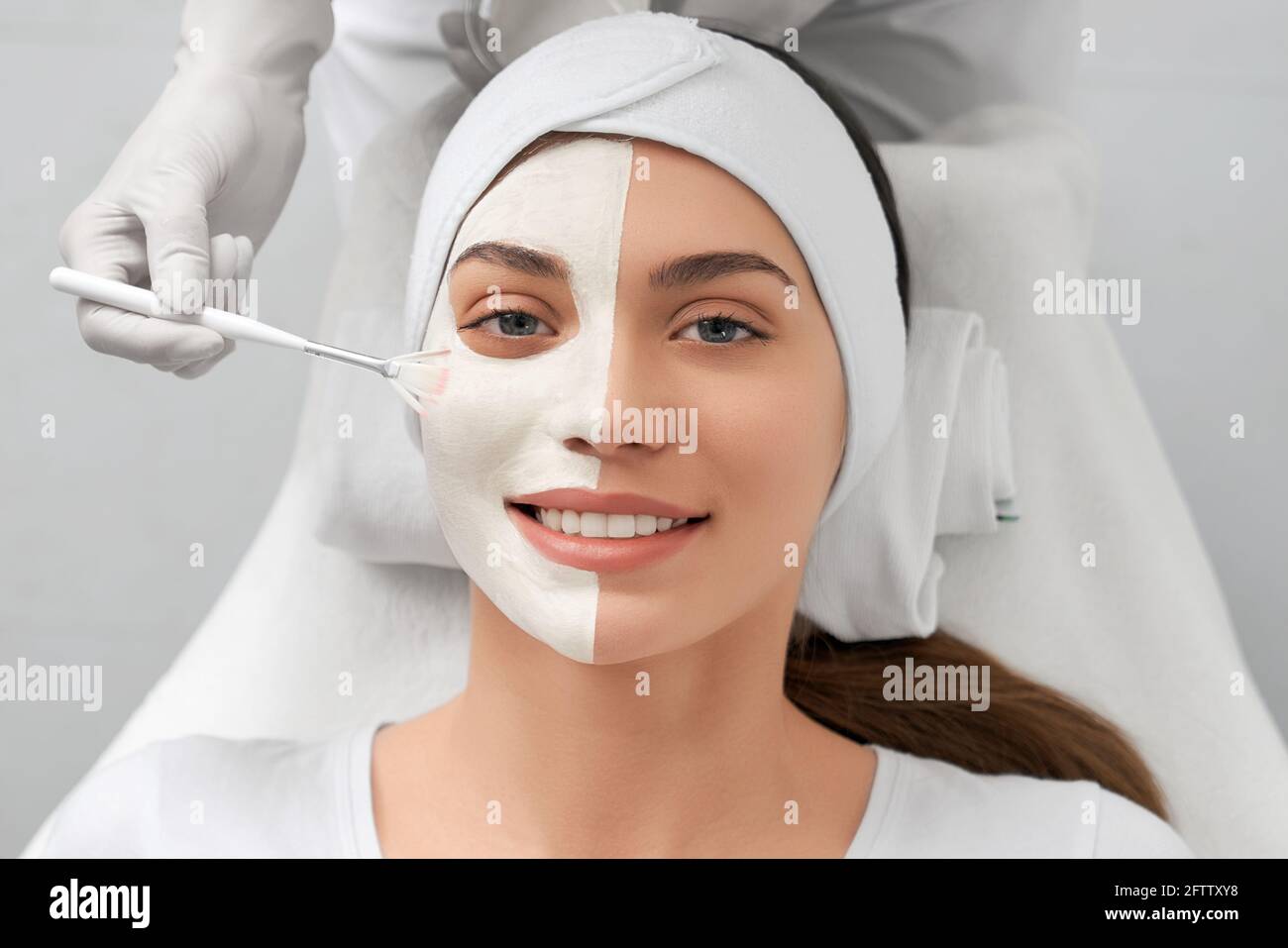Portrait of smiling young woman lying in beauty salon and beautician applying special white mask on face. Concept of procedure for rejuvenation skin or improvements. Stock Photo