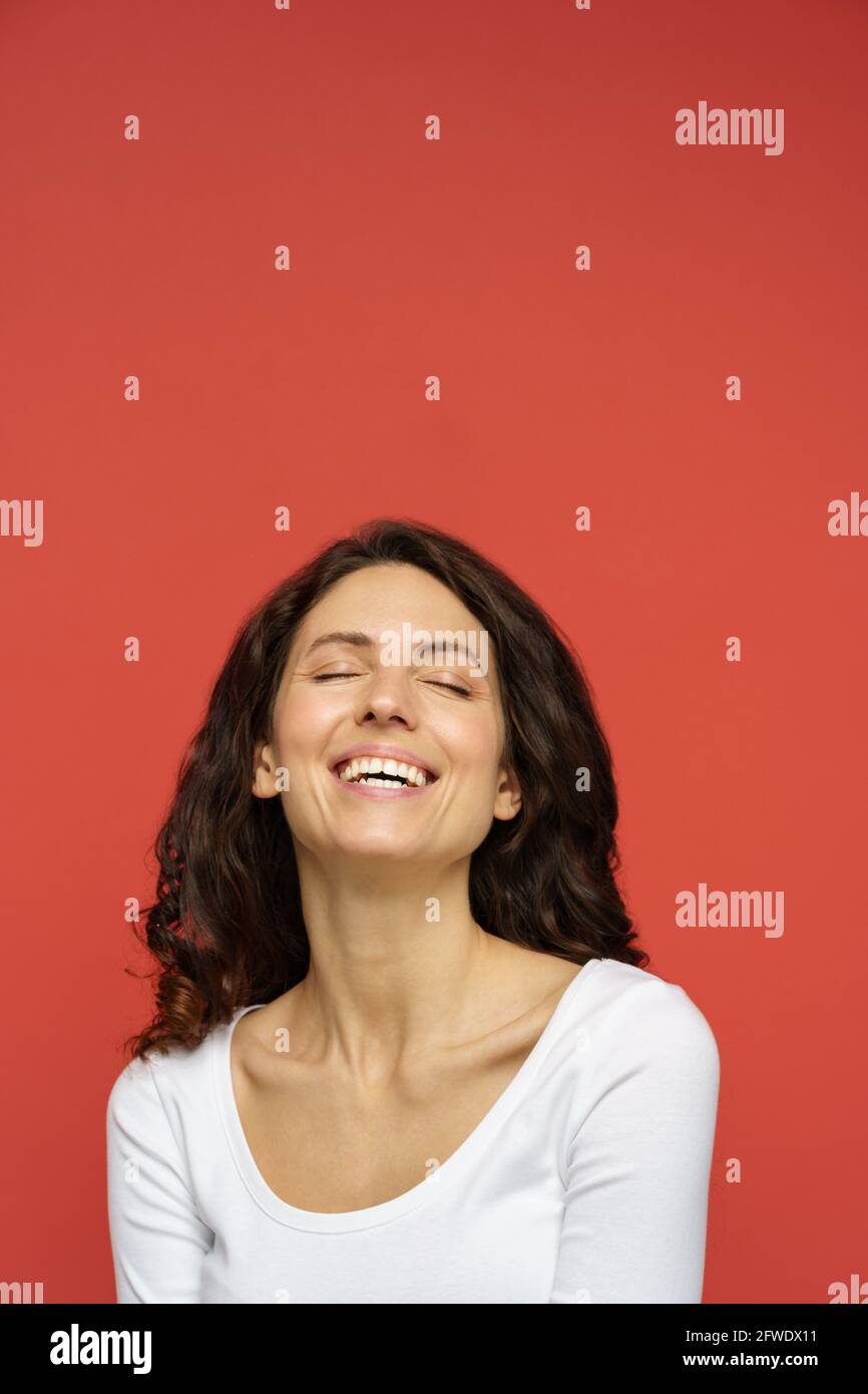 Portrait of carefree female in 30s with closed eyes, perfect skin, white smile on red background Stock Photo