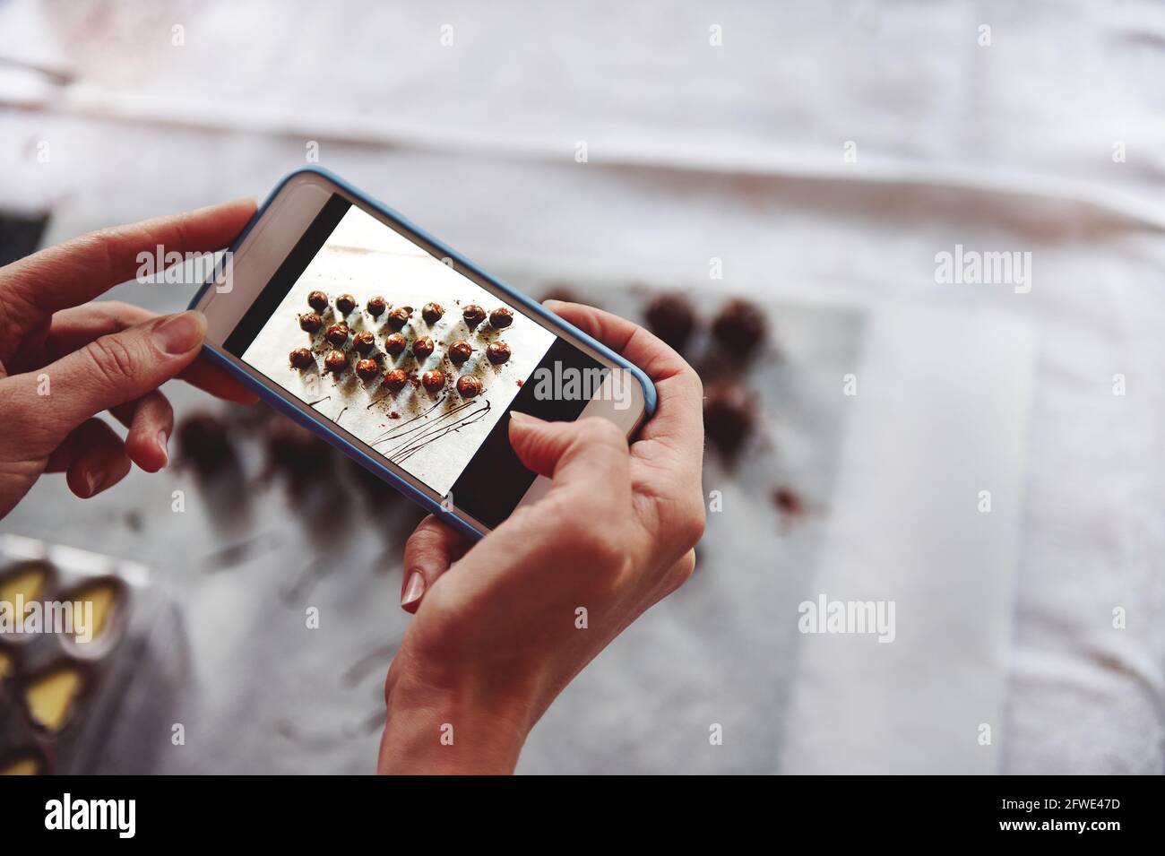 Closeup of hands holding mobile phone and making photography of delicious chocolate pralines on a white tablecloth. Mobile phone in live view regime. Stock Photo