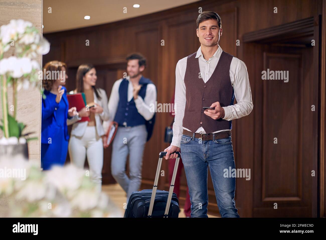 group of young business people, walking through the hallway. Selective focus on a male with cell phone, carrying wheeled suitcase. Stock Photo