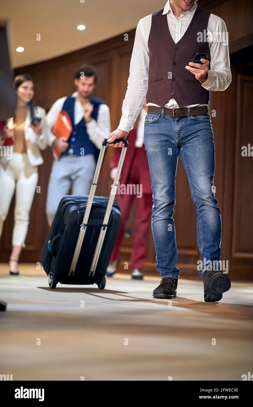 group of young adults, business people, walking through the hallway. Selective focus on a male with cell phone, carrying wheeled suitcase. Cropped ima Stock Photo