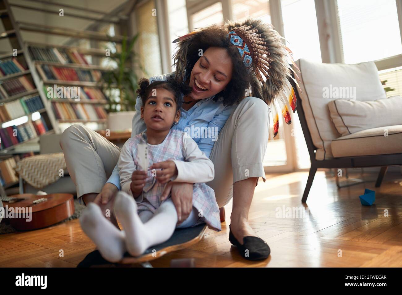 A young Mom with an indian headdress rides on the skateboard with her little daughter in a cheerful atmosphere at home. Family, together, love, playti Stock Photo