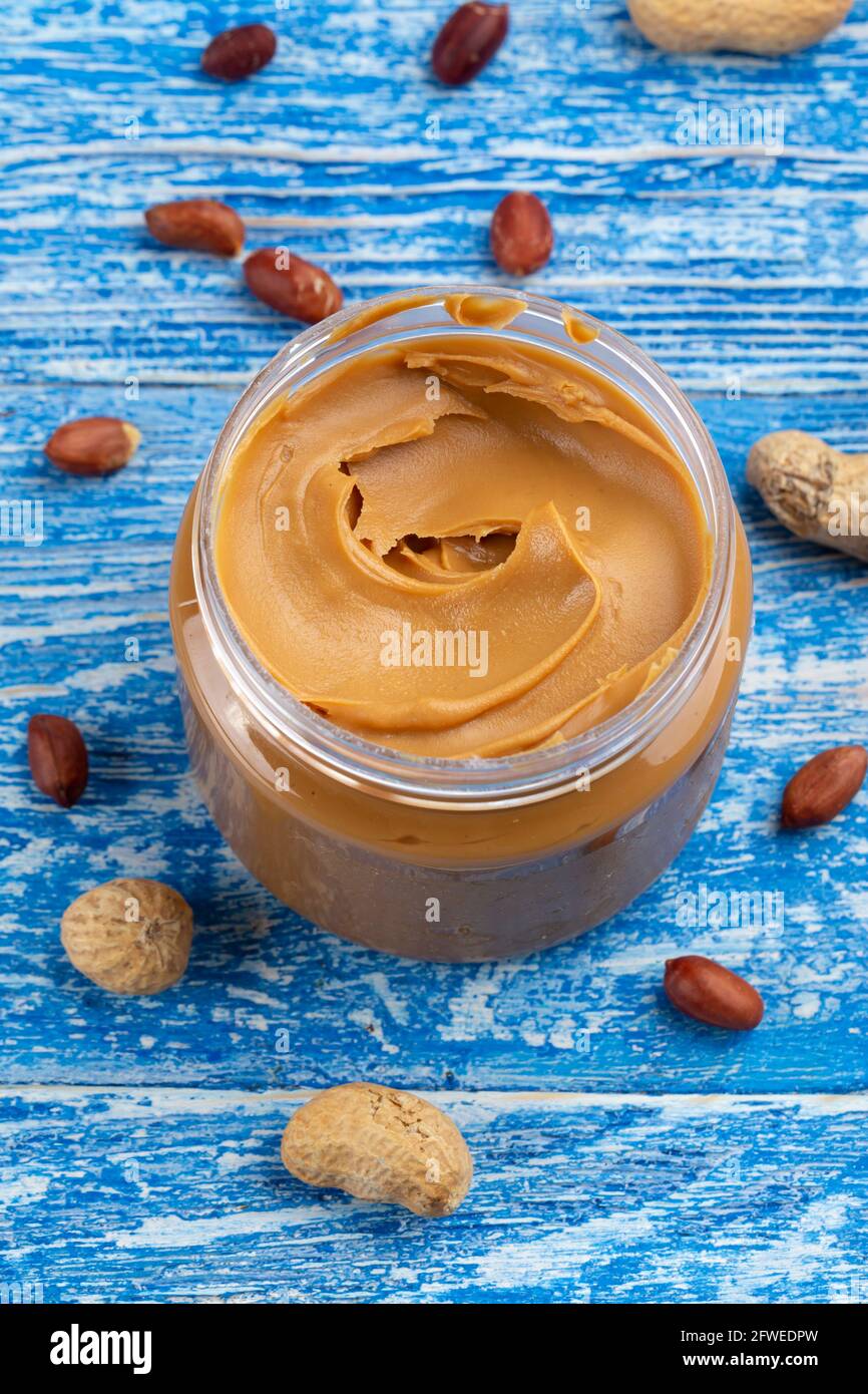 Peanut butter in an open jar and peanuts in the skin are scattered on the blue table. Space for