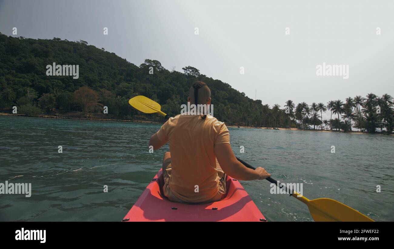 Man with sunglasses and hat rows pink plastic canoe along sea against green hilly islands with wild jungles. Traveling to tropical countries. Stock Photo