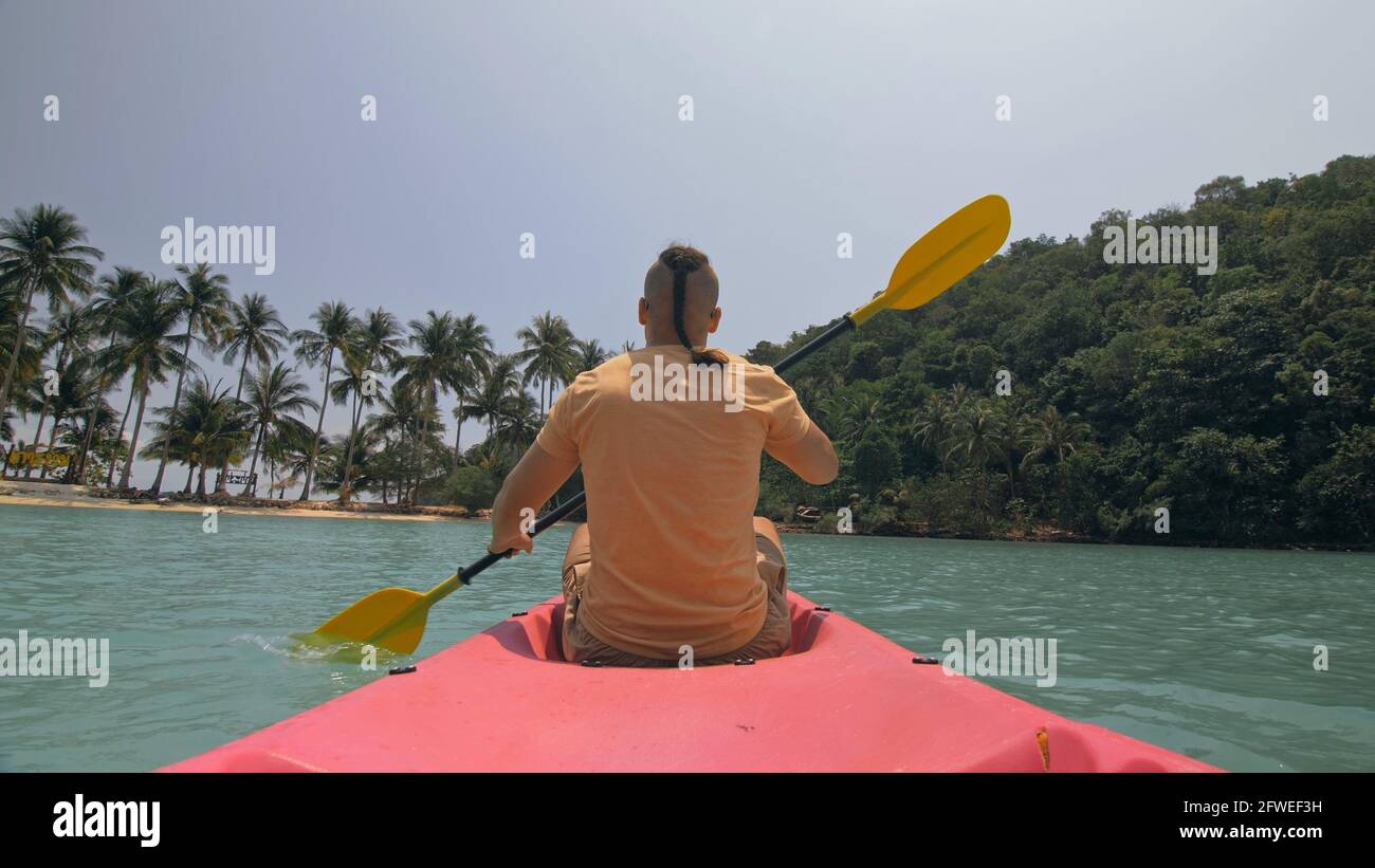 Man with sunglasses and hat rows pink plastic canoe along sea against green hilly islands with wild jungles. Traveling to tropical countries. Stock Photo