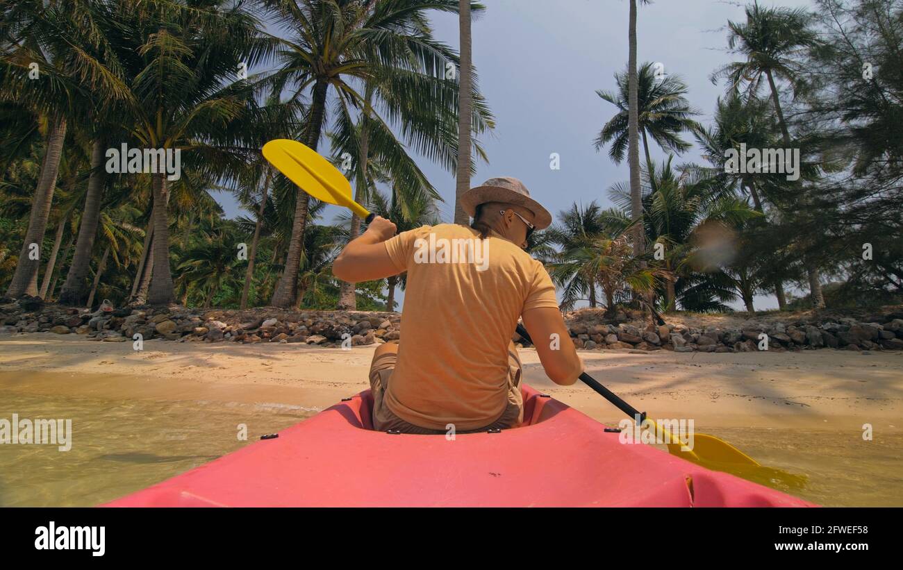 Man with sunglasses and hat rows pink plastic canoe along sea against green hilly islands with wild jungles. Traveling to tropical countries. Stock Photo