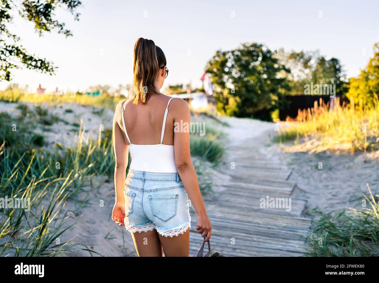 Summer tan on skin. Woman walking on boardwalk and leaving the beach. Stylish model wearing trendy jeans shorts. Hipster girl enjoying vacation. Stock Photo