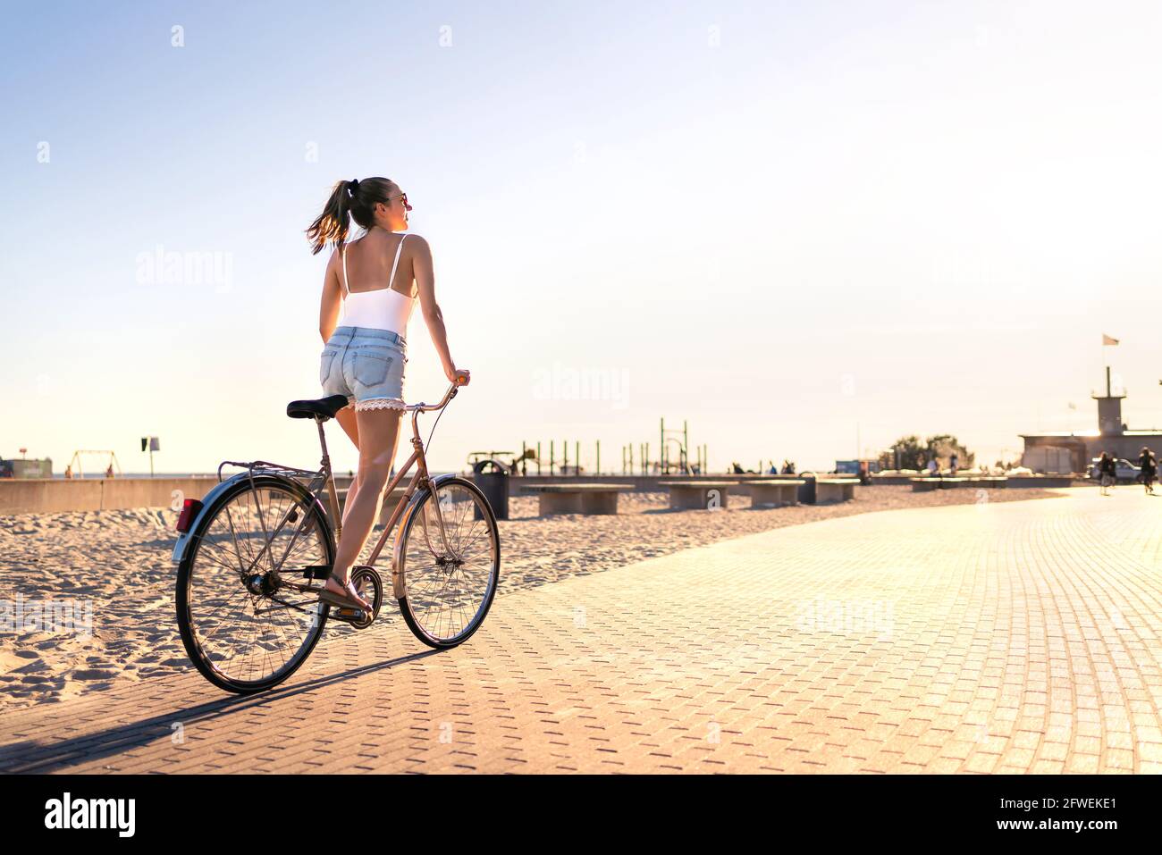 Bicycle fun on beach promenade. Happy woman riding bike on sunny summer boulevard. Seaside waterfront street for cycling. Stylish cyclist lady. Stock Photo