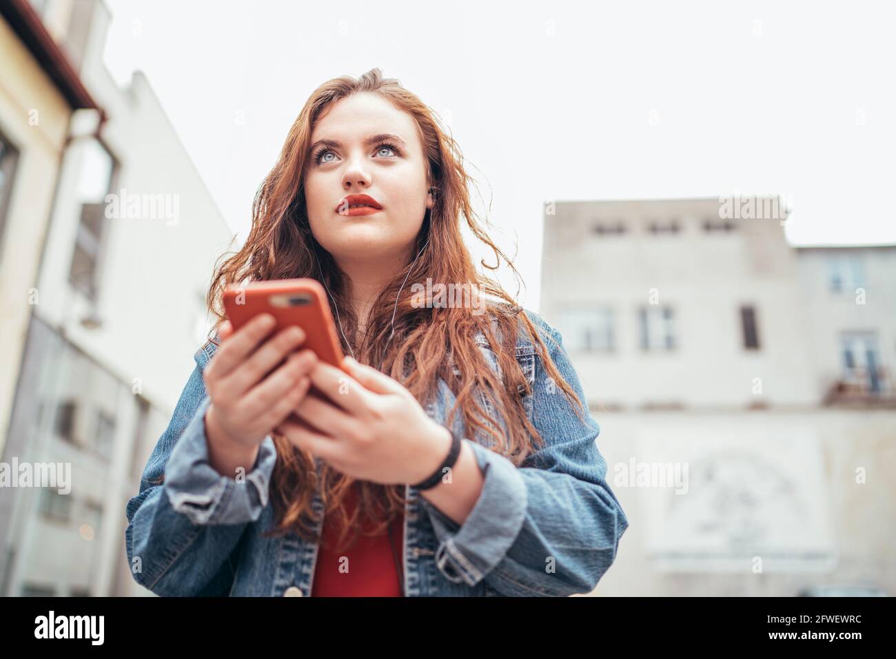 Curly red long hair caucasian teen girl walking on the street and using modern smartphone maps for city streets navigation. Modern people or technolog Stock Photo