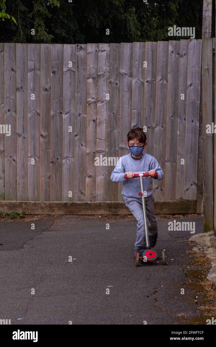 Full-length shot of a redhead boy wearing a face mask, riding a red scooter on a tar path surrounded by wood fences Stock Photo