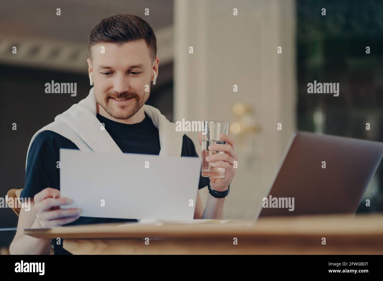 Smiling attractive male freelancer sitting in front of laptop holding glass of water Stock Photo