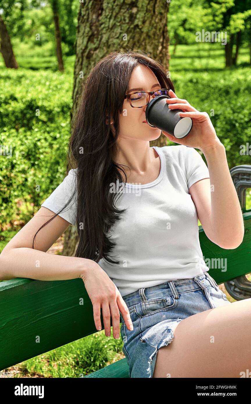 Close-up portrait young pretty woman drinking coffee from black paper cup sitting on a bench in the park. Stock Photo