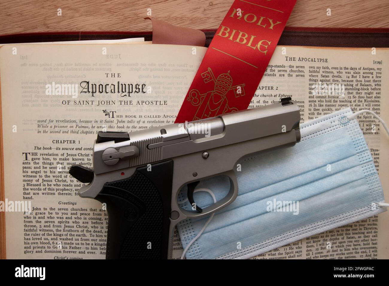 A gun resting on a bible open to the book of the Apocalypse with a Covid-19 protection mask and ammo Stock Photo