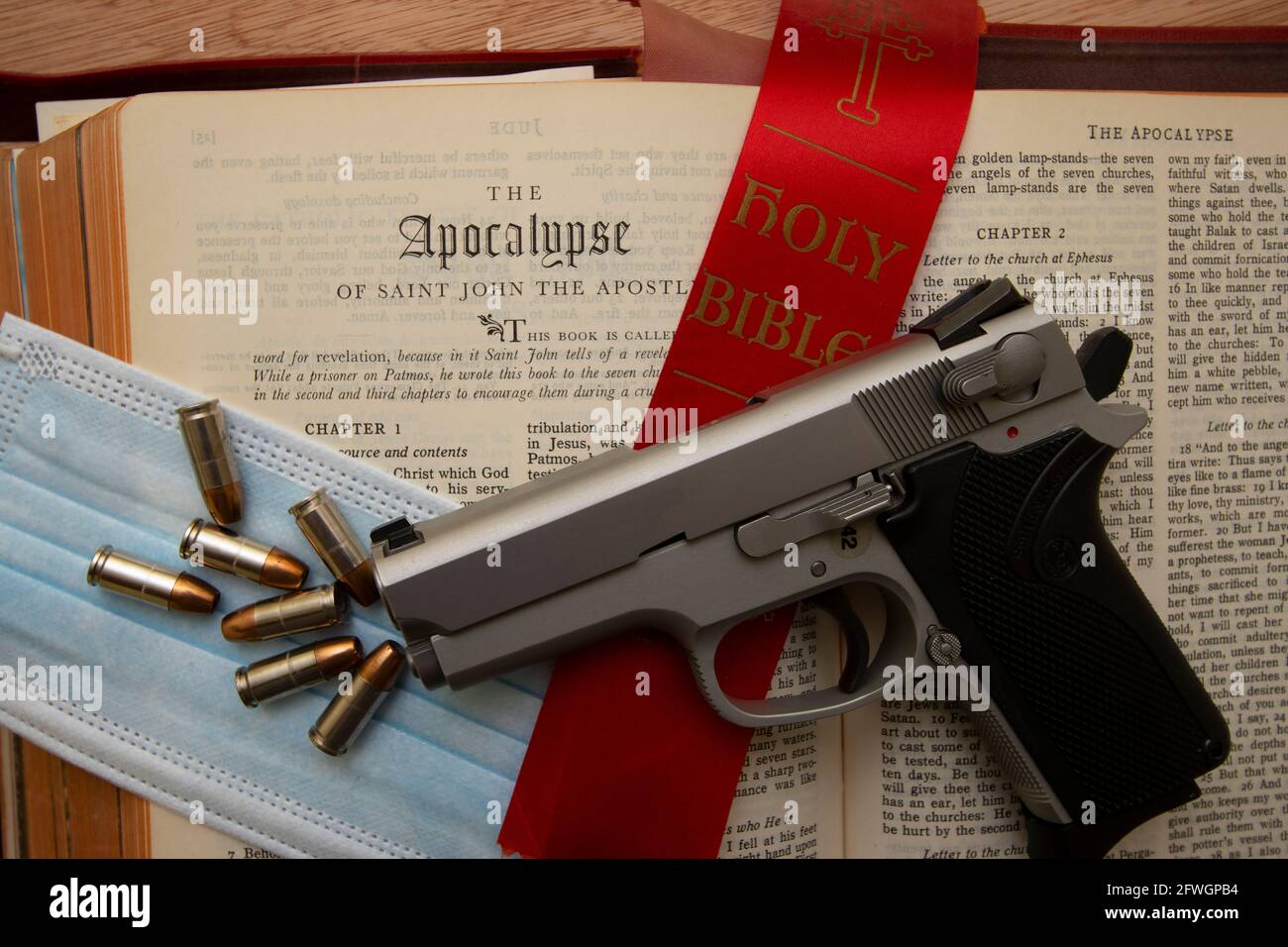 A gun resting on a bible open to the book of the Apocalypse with a Covid-19 protection mask and ammo Stock Photo