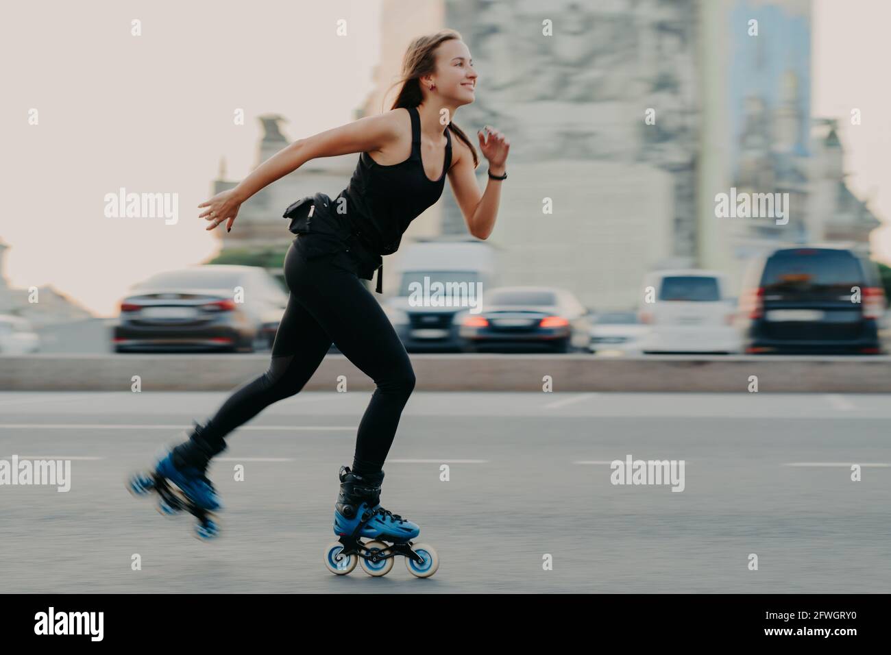 Slim healthy woman has recration time enjoys rolleblading on speed moves fast has cheerful expression wears black t shirt and leggings. Active rest Stock Photo