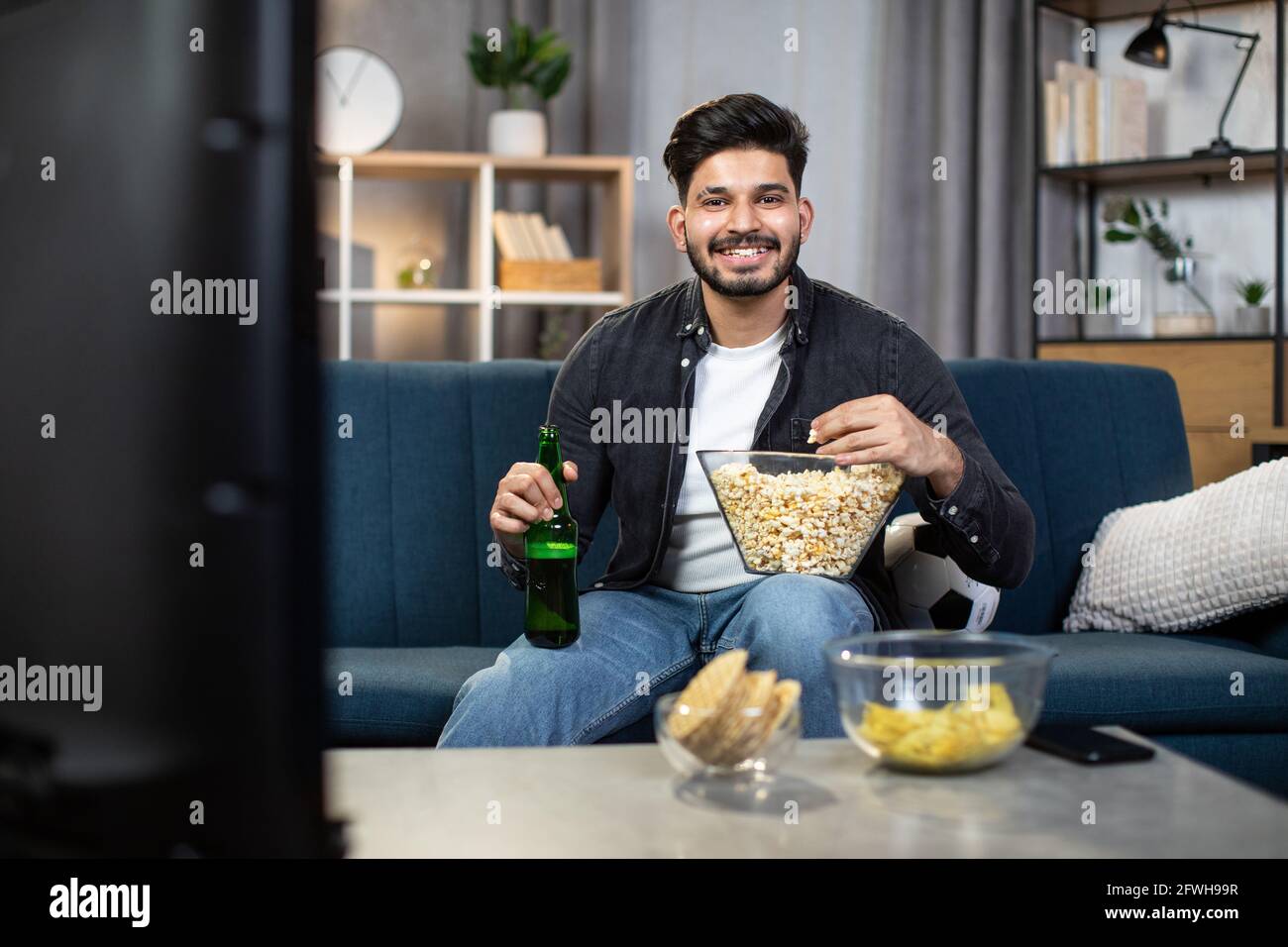 Positive indian guy in casual outfit enjoying tasty snacks and cold beer during football viewing on TV. Concept of people, leisure time and sport. Stock Photo