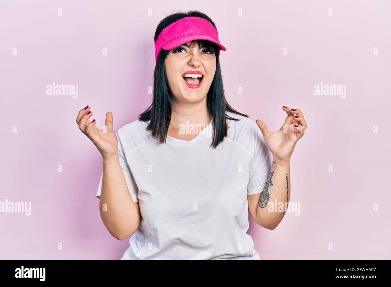 Young hispanic woman wearing sportswear and sun visor cap crazy and mad shouting and yelling with aggressive expression and arms raised. frustration c Stock Photo