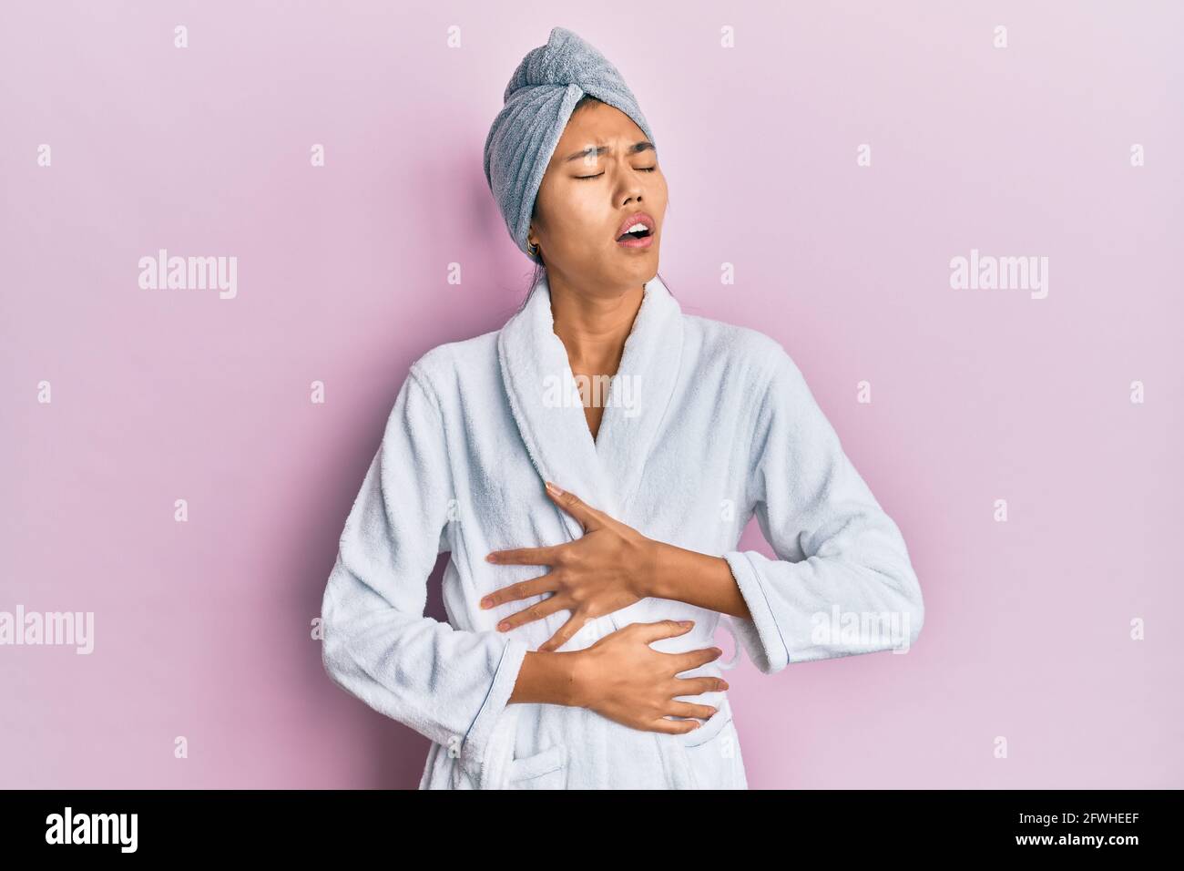 Young chinese woman wearing shower towel cap and bathrobe with hand on stomach because nausea
