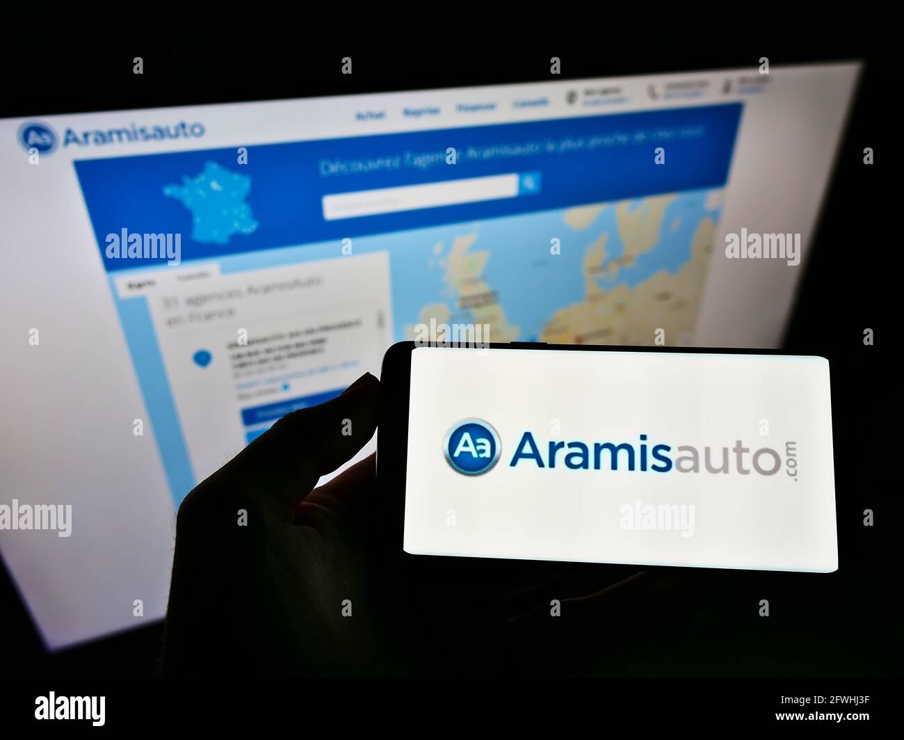 Person holding mobile phone with logo of French online car dealer Aramis SAS (Aramisauto) on screen in front of web page. Focus on cellphone display. Stock Photo