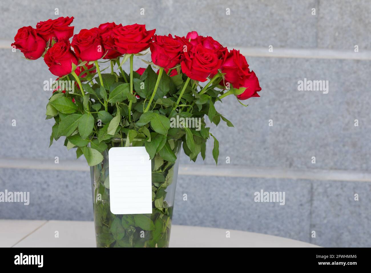 Bouquet of red roses in a vase and a white note for writing a greeting. Stock Photo