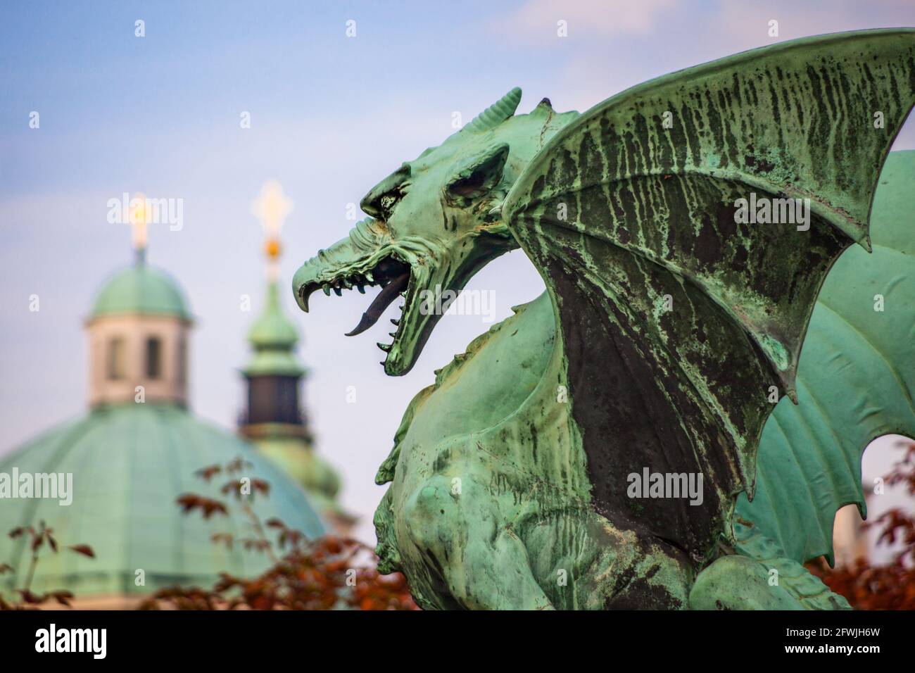 Bronze statue of the fabled Ljubljana Dragon, guarding the Dragon Bridge that crosses the Ljubljanica River, with the dome and spires of the Ljubljana Stock Photo