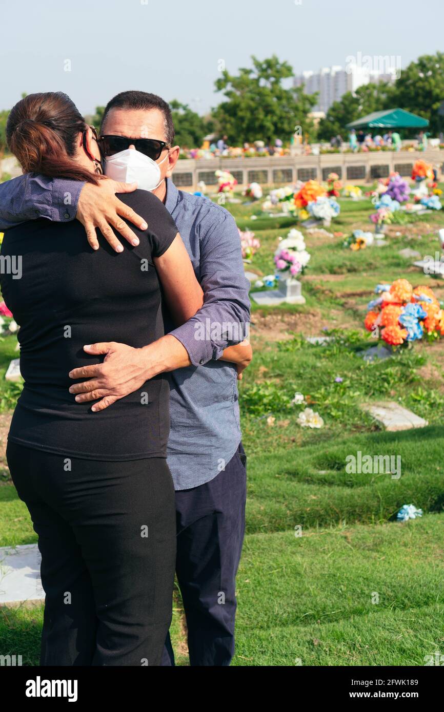 Husband with protective mask trying to comfort his wife in a cemetery. Tragic results of the coronavirus Stock Photo