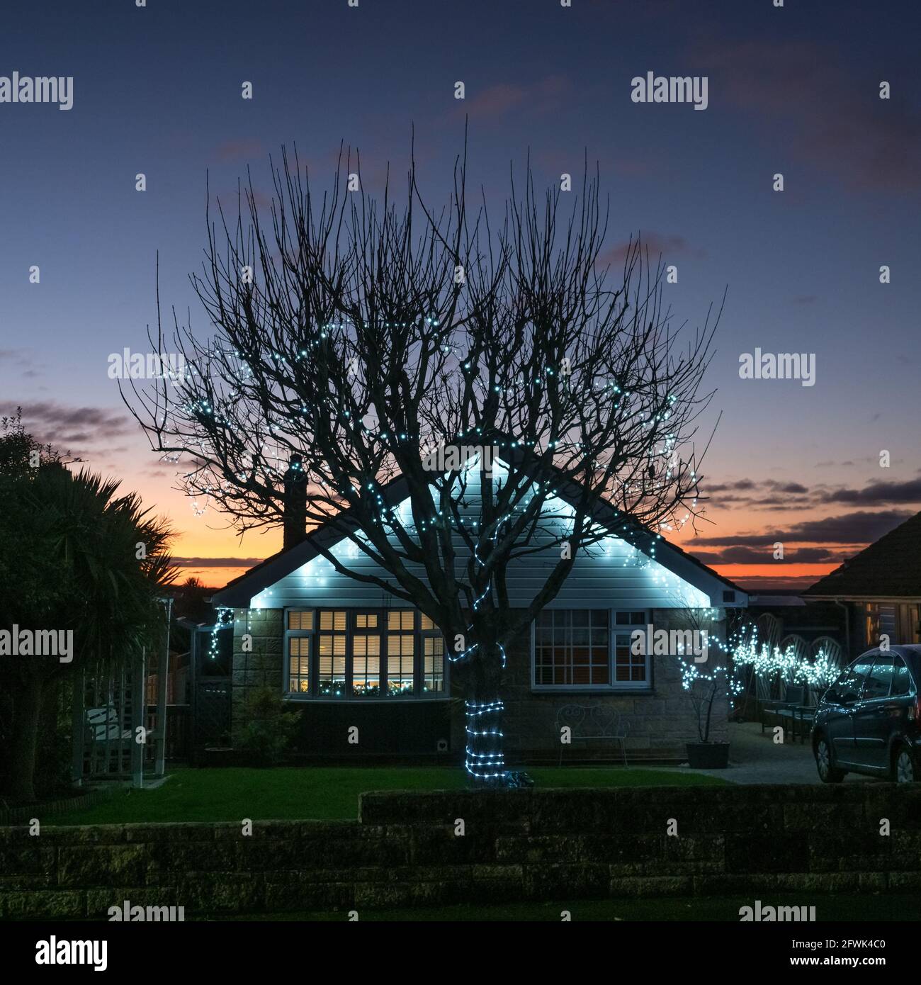 A front elevation view of blue Christmas lights in eaves of a bungalow house home and hanging in tree silhousette with sunset in blue sky as backdrop Stock Photo