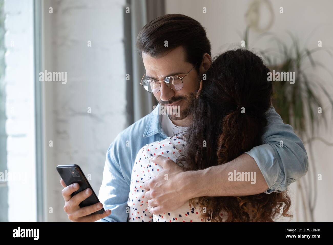 Dishonest young man using phone while cuddling wife. Stock Photo