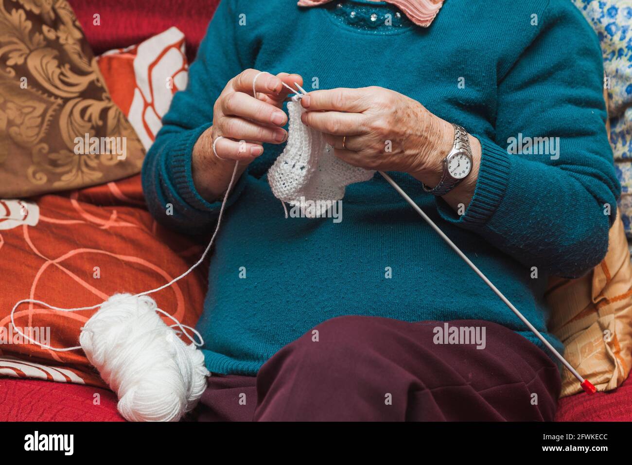 The hands of an elderly lady who is sitting on a sofa knitting a white woolen garment with long needles. She wears maroon pants and green sweater, on Stock Photo