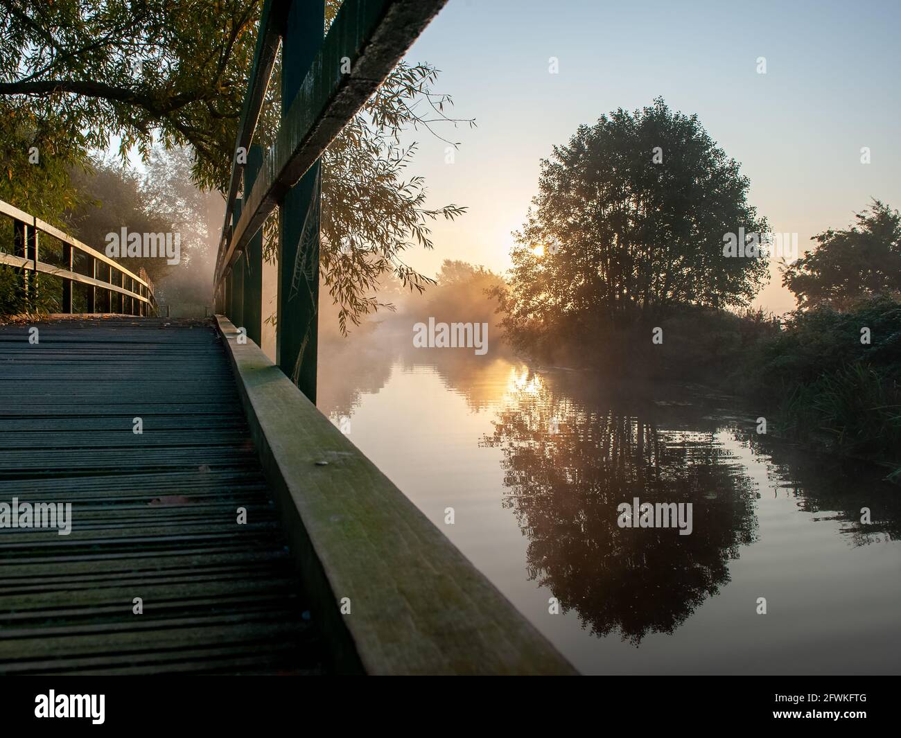 Wooden footbridge over river at sunrise with layer of mist and trees reflected in water beneath a clear sky with still waters Stock Photo