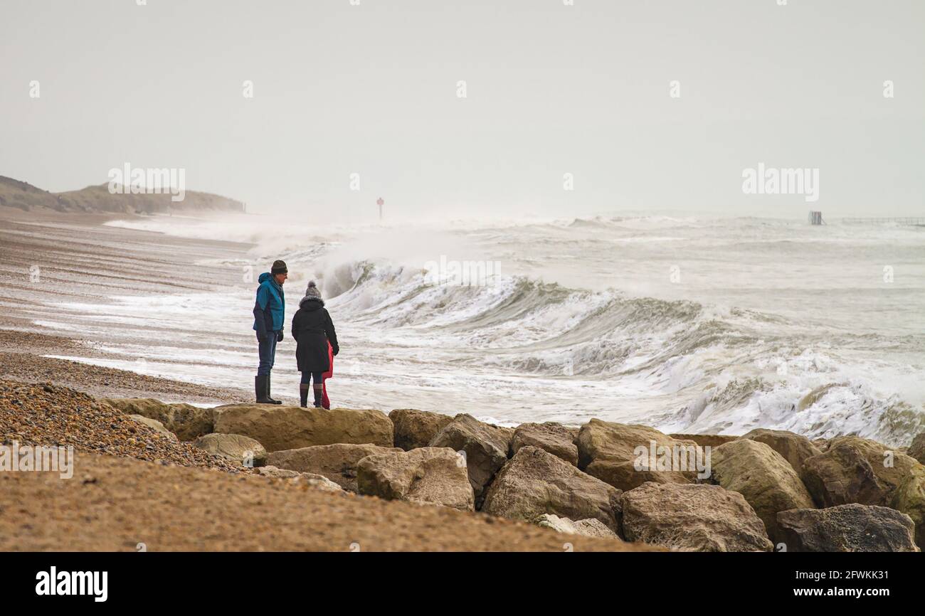 Family Wrapped Up Against The Cold Standing On A Groyne Made Of Rocks Watching The Waves During A Winter Storm On Hengistbury Head Beach UK Stock Photo